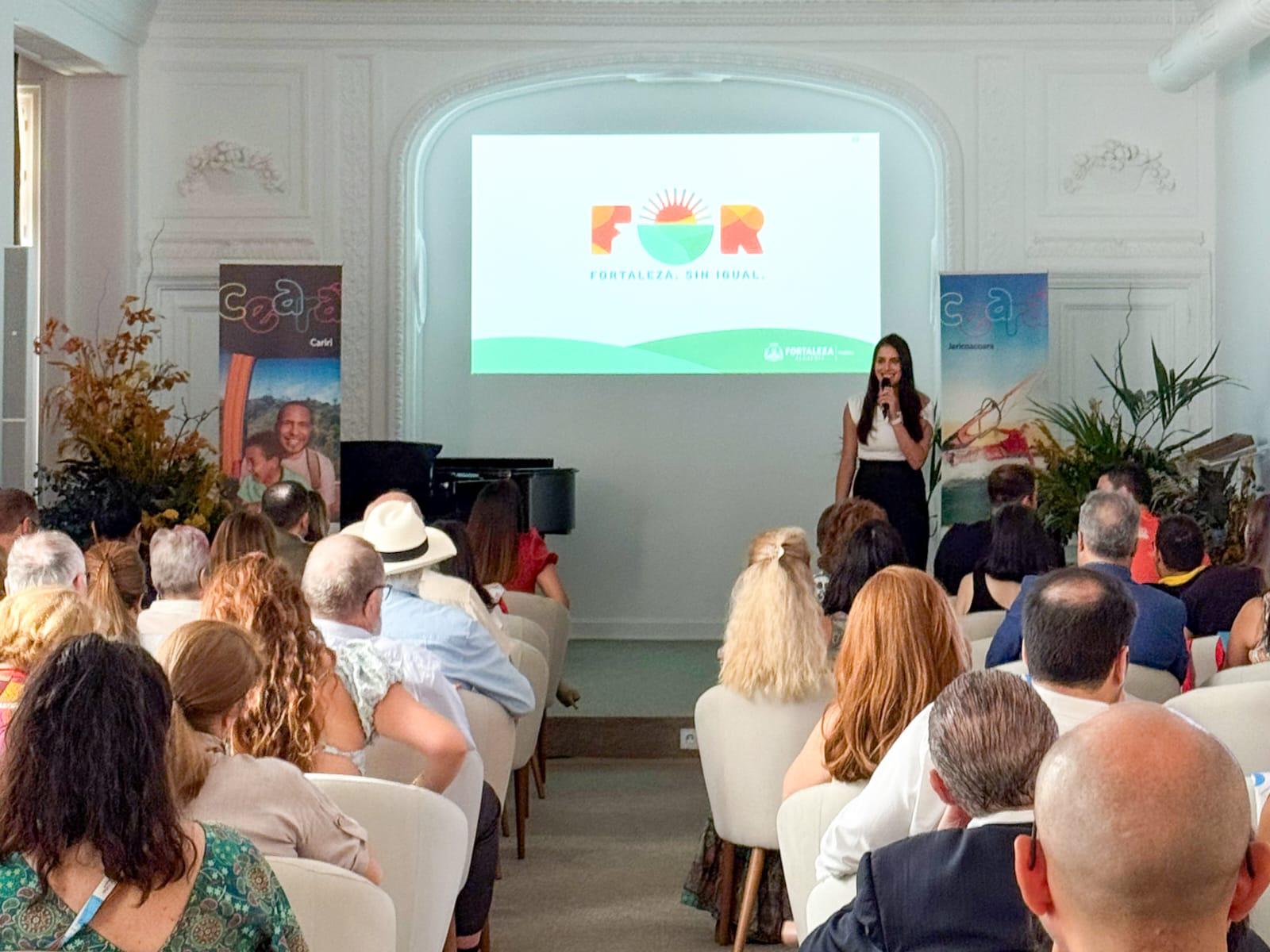 a foto traz uma imagem aberta de um auditório com pessoas sentadas em cadeiras